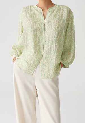 Personne portant un chemisier boutonné à manches longues à motif vert clair et un pantalon ample blanc cassé, les mains dans les poches, sur un fond uni.