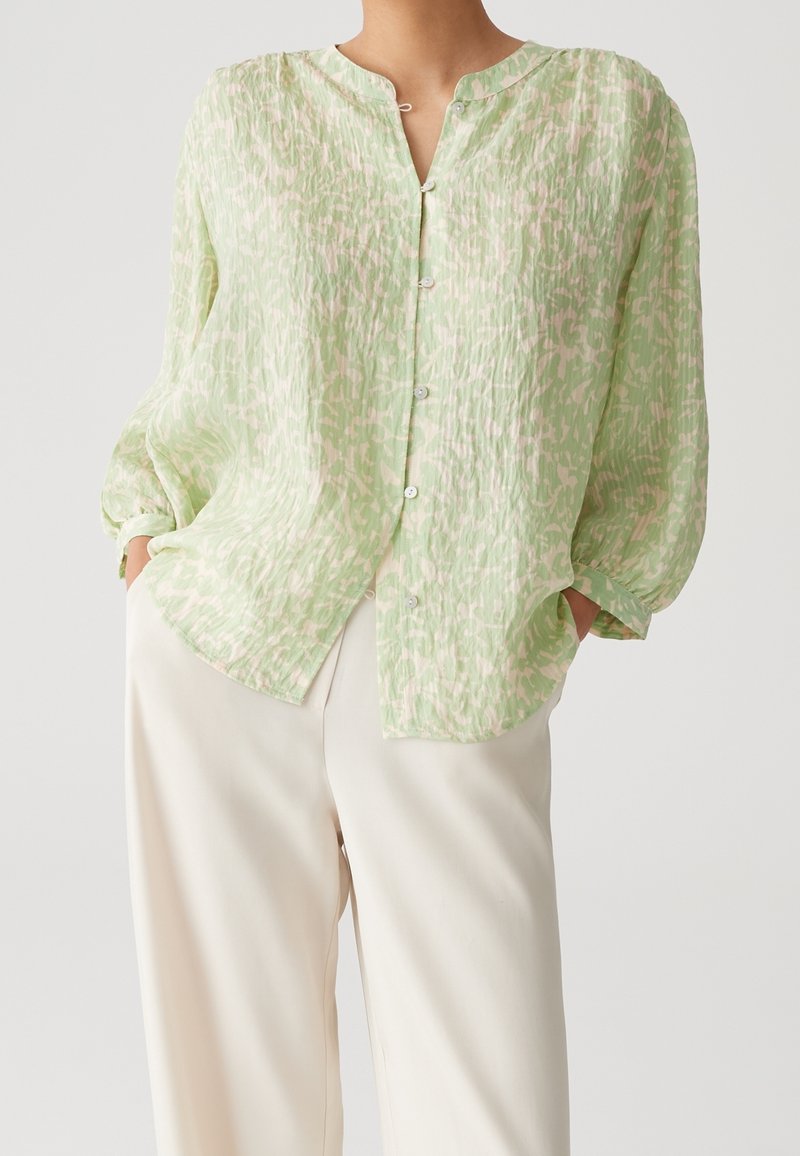 Personne portant un chemisier boutonné à manches longues à motif vert clair et un pantalon ample blanc cassé, les mains dans les poches, sur un fond uni.