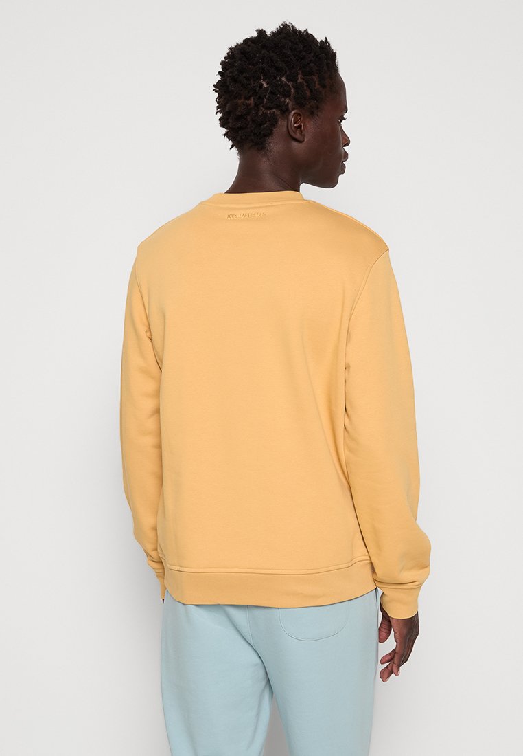 Gelbes Sweatshirt mit Rundhalsausschnitt, gerippten Bündchen und Saum. Glatte Textur mit schlichtem Design, getragen mit hellblauen Hosen.