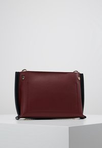 Bolso de piel burdeos con detalles en negro, forma rectangular, textura suave y herrajes de metal; cuenta con un bolsillo frontal y asas superiores.