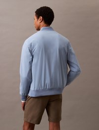 Giacca bomber azzurro chiaro con colletto e polsini a costine, vita elasticizzata e texture liscia, abbinata a pantaloni corti marroni.