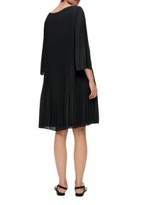 Svart plisserad klänning med trekvartslånga volangärmar, med rundad halsringning och flytande fåll, tillsammans med svarta sandaler.