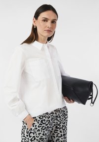 Biała koszula zapinana na guziki z dwiema kieszeniami na piersi, zestawiona z jasnoszarymi spodniami w czarny nadruk z lampartem. Modelka trzyma czarną torebkę kopertową.
