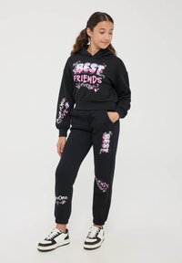 Completo di felpa nera con cappuccio e pantaloni della tuta. La felpa presenta una grafica testuale rosa "MIGLIORI AMICI" e grafiche sui gomiti. I pantaloni hanno grafiche simili e polsini a coste.