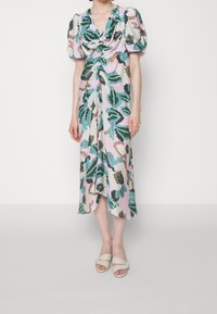 Mujer vistiendo un vestido midi con mangas abullonadas, un frente fruncido y patrones florales abstractos en verde, negro, marrón y azul, combinado con sandalias blancas.
