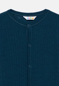 Tmavomodrý rebrovaný detský overal s okrúhlym výstrihom a tromi patentkami na prednej strane, s farebnou značkovou etiketu vo vnútri goliera.