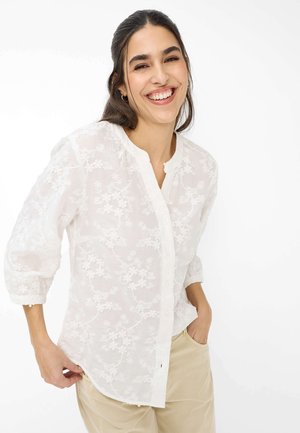 Lächelnde Frau mit dunklen Haaren trägt eine weiße Bluse mit floraler Stickerei und 3/4-Ärmeln sowie beige Hosen, die Hände in den Taschen, steht vor weißem Hintergrund.