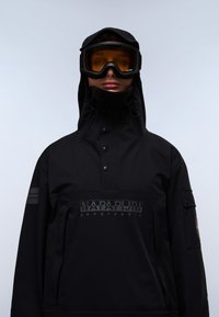 Persona che indossa una giacca nera con cappuccio e colletto alto, e occhiali da sci con lenti arancioni su uno sfondo chiaro e semplice.
