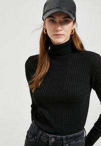 Schwarzer gerippter Rollkragenpullover mit langen Ärmeln, aus weichem Stoff, kombiniert mit dunklen Jeans und einer schwarzen Lederkappe.
