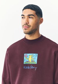 Vinröd sweatshirt med en grafisk design av en DJ med gula och blå accenter. Texten nedanför grafiken lyder "Keith Haring™".