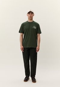 Zelené bavlněné tričko s logem, kombinované s tmavými džínovými kalhotami a hnědými botami. Model nosí světle béžovou čepici a stojí na neutrálním pozadí.