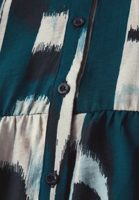 Abito a camicia con motivo astratto bianco, blu e nero. Presenta una vita arricchita e bottoni in plastica nera. Tessuto liscio e traspirante.