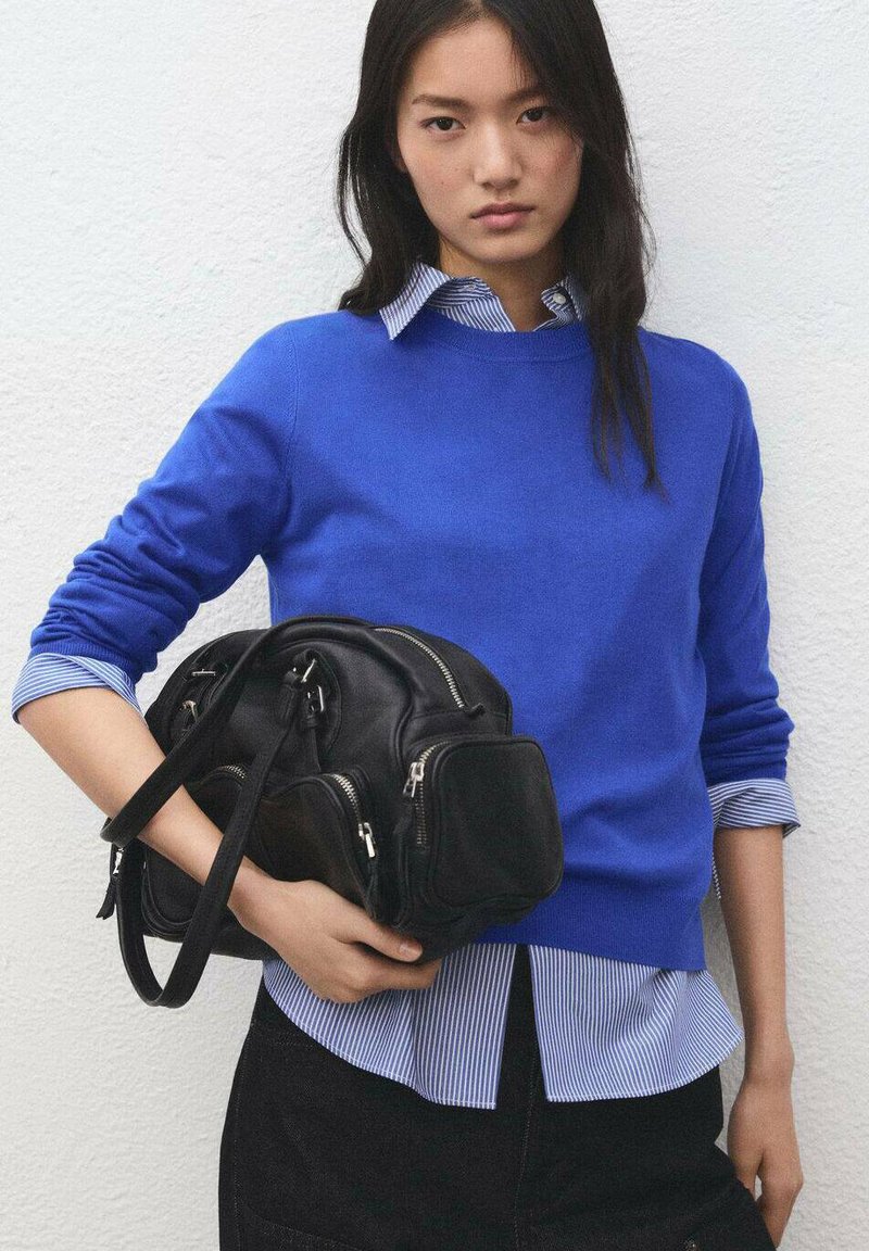 Maglione blu reale sopra una camicia a righe blu e bianche, abbinato a una borsa in pelle nera con molteplici cerniere e un design strutturato.