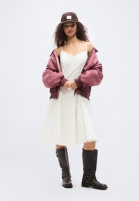 Γυναίκα με λευκό φόρεμα, φαρδιά μωβ ζακέτα που γλιστράει από τους ώμους, καφέ καπέλο και καφέ σκούρα δερμάτινα μποτάκια μέχρι το γόνατο, στέκεται μπροστά σε απλό φόντο.