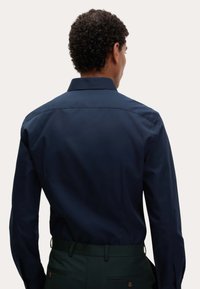 Navyblauw button-up shirt met een klassieke kraag en een gladde textuur, gezien vanuit de achterkant, gecombineerd met groene broeken.