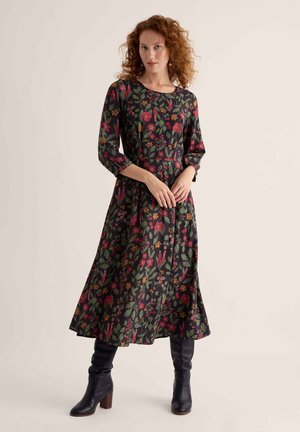 Femme aux cheveux roux bouclés portant une robe sombre à manches longues avec un motif floral rouge et vert et des bottes noires montantes.