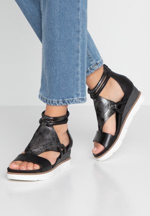 Czarne sandały z metalicznym srebrem o otwartym fasonie, z paskami na kostkach, teksturowanym wierzchem i białą podeszwą na koturnie. Na zdjęciu widoczne są również dżinsy.