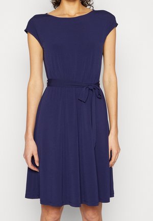 Femme portant une robe bleu marine mi-longue avec des manches courtes et une ceinture nouée à la taille, debout devant un fond clair uni.