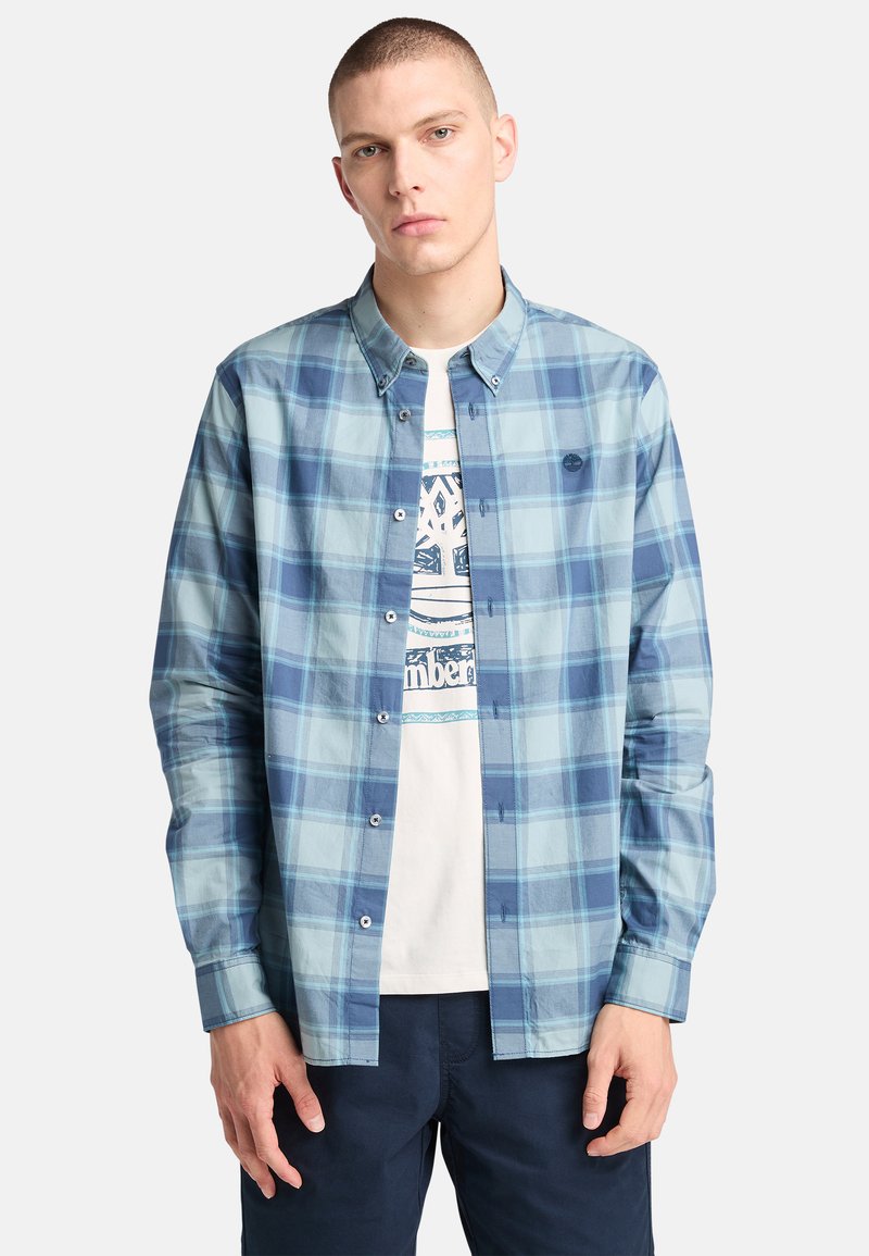 Camicia a quadri blu chiaro con colletto button-down, maniche lunghe e bottoni bianchi, indossata sopra una t-shirt bianca con grafica.