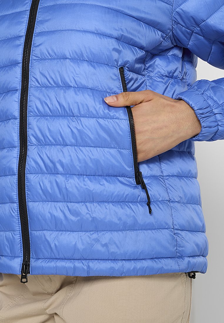 Eine Hand teilweise in der Reißverschlusstasche einer blauen gesteppte Jacke, die mit beigen Hosen getragen wird.