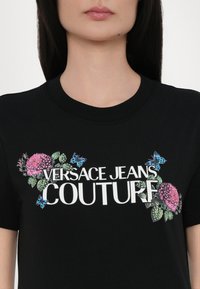 Juodas medvilnės marškinėliai su užrašu "VERSACE JEANS COUTURE" baltos spalvos, papuošti spalvingomis gėlių ir drugelių grafikomis kairėje pusėje.