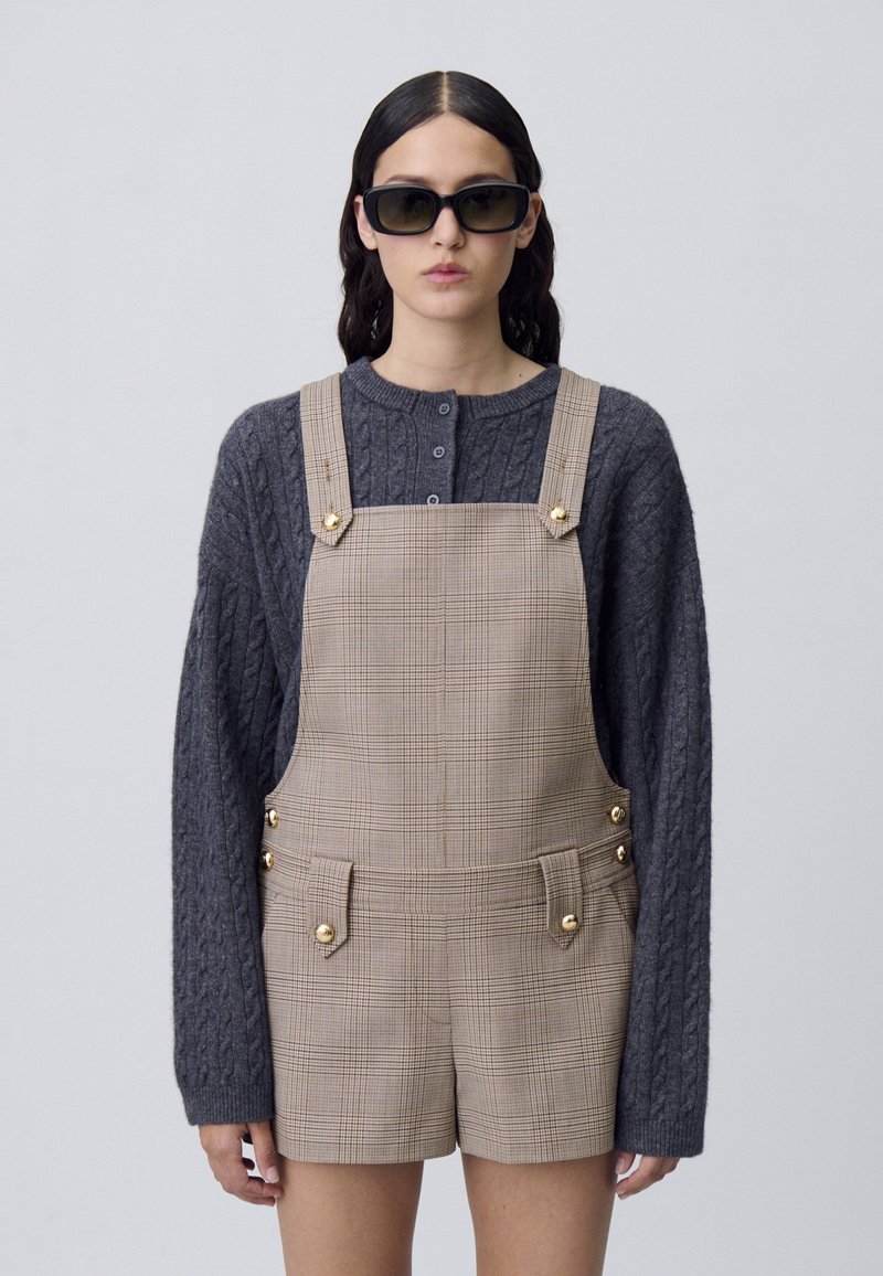 Suéter gris de punto grueso en talla grande, combinado con tirantes cortos de cuadros en color beige que presentan detalles de botones dorados y bolsillos delanteros. Lleva puestas gafas de sol negras.