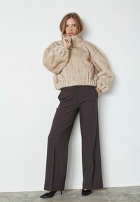 Giacca bomber in pelliccia sintetica beige con collo alto e orlo arricciato, abbinata a pantaloni a gamba larga marrone scuro e tacchi a punta neri.