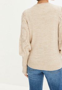 Kvinna som bär en beige stickad tröja med spets- och bollar-mönster på ärmarna och blå denimjeans, sedd bakifrån.