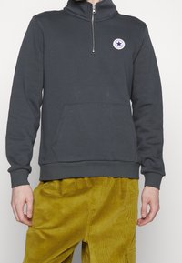 Sudadera gris de punto con un cuello de media cremallera, bolsillo canguro frontal y un parche de logo circular con una estrella. Textura de tela suave.