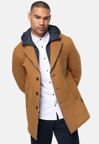 Brun kappa med marinblå huva, med skräddarsydd design, knapplåsning och sidofickor. Bärs över en vit skjorta och mörka jeans.