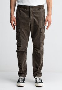 Bruine cargobroek met een elastische tailleband, meerdere zijzakken en een ontspannen pasvorm. Gemaakt van een zachte, duurzame stof. Draagt een paar zwarte sneakers.