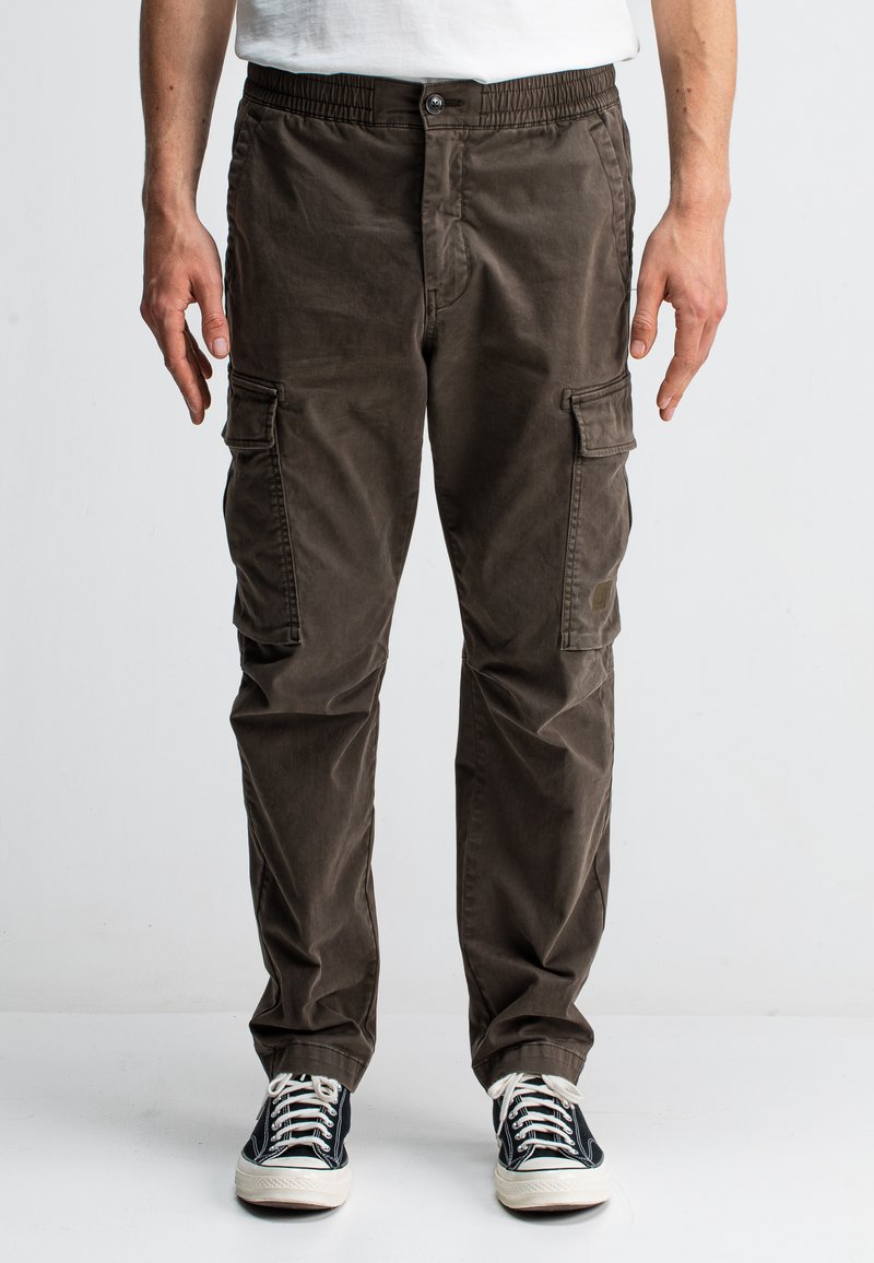 Bruine cargobroek met een elastische tailleband, meerdere zijzakken en een ontspannen pasvorm. Gemaakt van een zachte, duurzame stof. Draagt een paar zwarte sneakers.