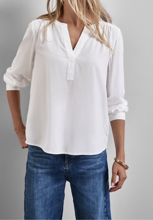 Vrouw draagt een witte blouse met lange mouwen en een V-hals en blauwe spijkerbroek, met los blond haar en een gouden armband om de rechterpols.