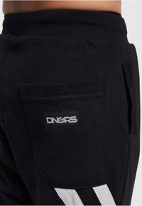 Schwarze Jogginghosen verfügen über eine Gesäßtasche mit einem rechteckigen Patch mit der Aufschrift "DNGRS". Sie haben weiße Seitestreifen und einen gerippten Bund.