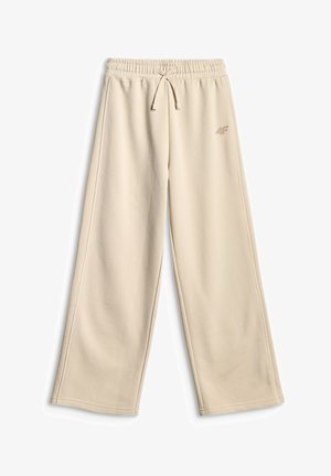 Jeune fille aux longs cheveux bruns vêtue d'un sweat à capuche beige et d'un pantalon assorti, debout les mains dans les poches sur un fond uni.