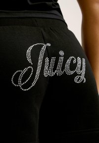 Fekete sportnadrág, amelyen csillogó ezüst strasszdesign található, hátul "Juicy" felirattal, puha anyagból készült, testhez simuló fazonnal.