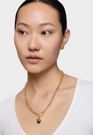 Kobieta o ciemnych włosach nosi złote kolczyki w kształcie serca oraz masywny złoty łańcuszek z małym brązowym wisiorkiem w kształcie serca, biała bluzka.