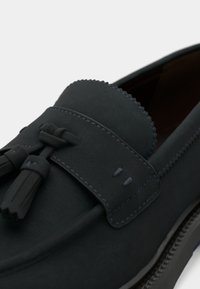 Sorte loafers i ruskind med flad sål, dekorativ kvast, kontrastfarvet blå syning og en rundet tå med en glat tekstur.