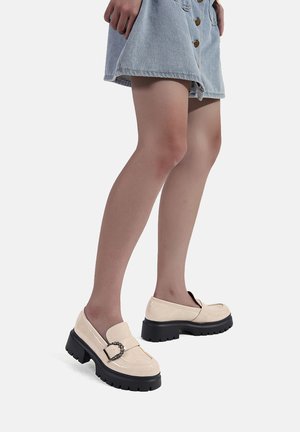 Mocassini in suede beige con una finitura lucida, caratterizzati da un accento a catena e una suola nera spessa. Indossati con una gonna di jeans, mostrando un profilo laterale.
