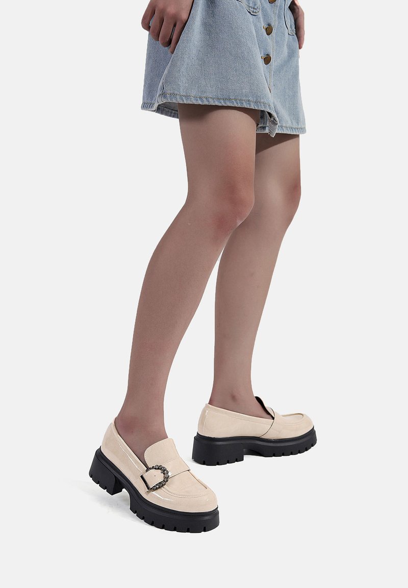 Mocassini in suede beige con una finitura lucida, caratterizzati da un accento a catena e una suola nera spessa. Indossati con una gonna di jeans, mostrando un profilo laterale.