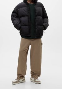 Sort dunjakke med vandret polstring, kombineret med en mørkegrøn skjorte og beige bukser med vide ben. Brune og hvide sneakers fuldender outfittet.