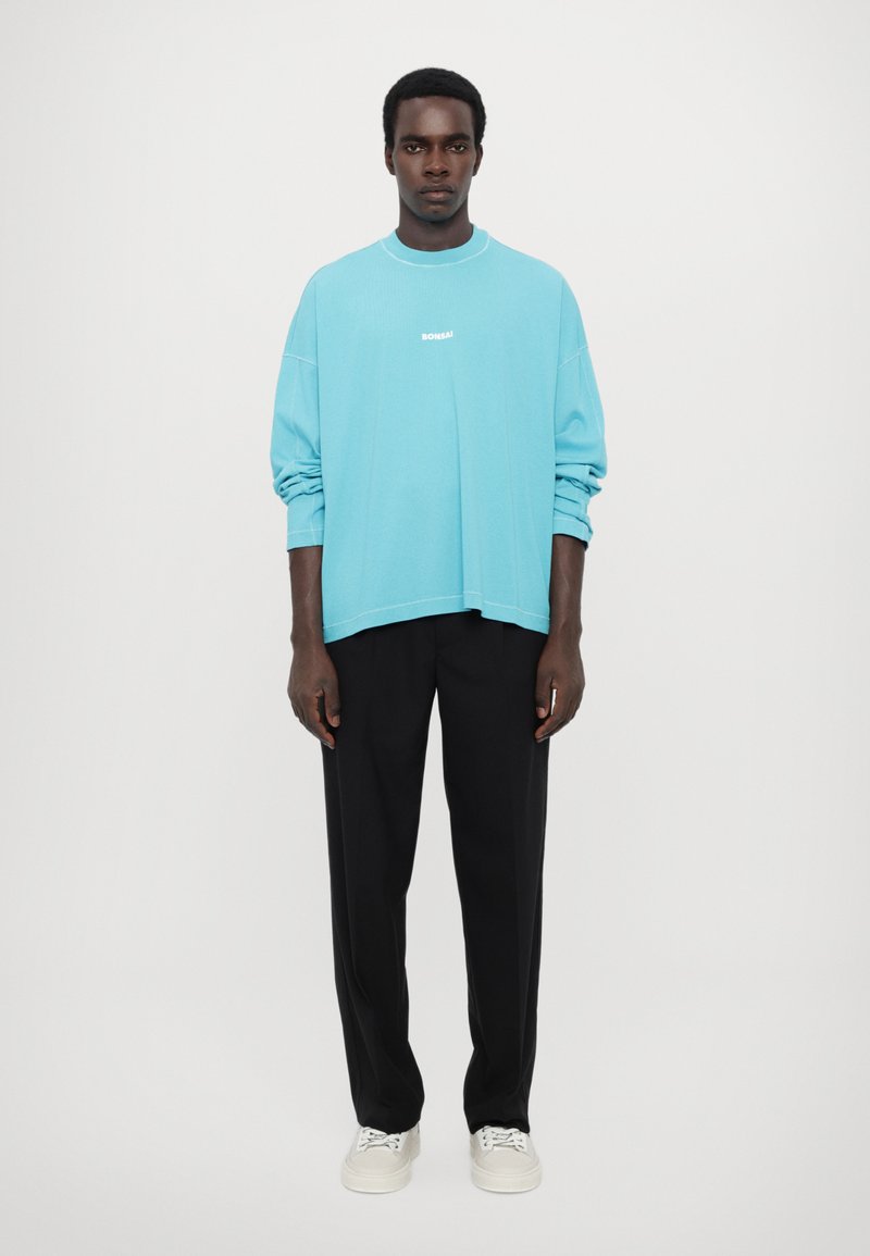 Light blue oversized long-sleeve shirt with white logo, paired with black trousers and white sneakers, standing against a plain background.