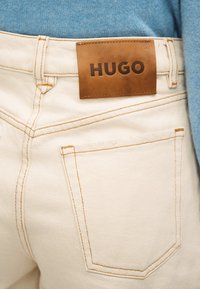 Κοντινό πλάνο πίσω τσέπης μπεζ τζιν με καφέ δερμάτινο λογότυπο με την επιγραφή "HUGO" πάνω από την τσέπη, φορεμένη με ανοιχτό μπλε τοπ.