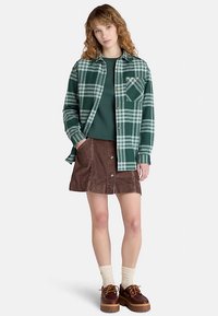 Camicia a quadri verde con bottoni e taschino sul petto sopra un maglione verde scuro, abbinata a una gonna di velluto a coste marrone. La modella indossa calzini crema e scarpe marroni.