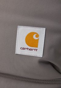 Surface en tissu gris avec un patch logo Carhartt en blanc, un emblème orange et un texte rouge. La texture semble solide et tissée.