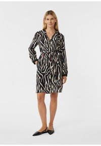 Donna che indossa un vestito zebra a metà coscia, nero e beige, con maniche lunghe e scarpe basse nere a punta, in piedi con una mano in tasca.