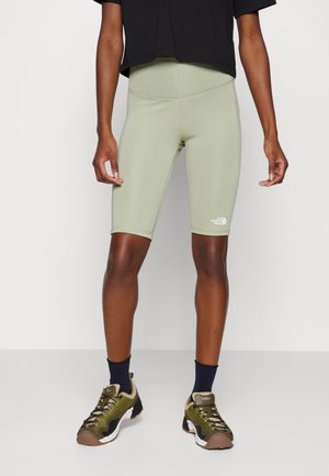 Persoon die lichtgroene sportshorts tot halverwege de dijen draagt met het The North Face-logo, een zwart cropped topje, zwarte sokken en groene wandelschoenen, staand.