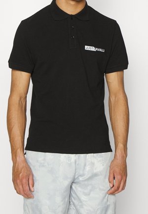 Mann trägt ein schwarzes Poloshirt mit dem "Just Cavalli"-Logo auf der Brust und hellfarbene, strukturierte Shorts, steht vor weißem Hintergrund.