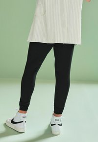 Colanți negri asortați cu un top alb cu dungi subțiri și adidași înalți albi cu accente negre. Fundalul este de un verde moale.