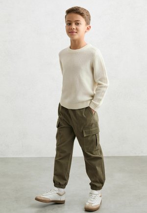 Krémový pletený sveter s rebrovanými manžetami, olivové cargo nohavice so bočnými vreckami a biele tenisky so sivými akcentmi. Jednoduché, ležérne oblečenie.